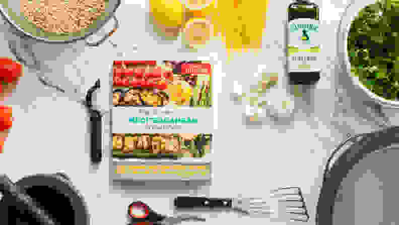 A book sits on a kitchen counter, surrounded by lemons, olive oil, garlic, and various tools. The cover reads, 