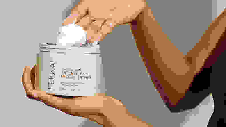 A person holding a gray jar and scooping out a white cream with their other hand.
