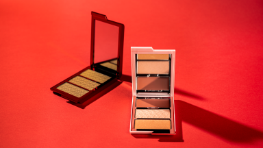 Two open makeup palettes, one in a bronze color and another in a light pink, sit arrayed against a red-orange background.