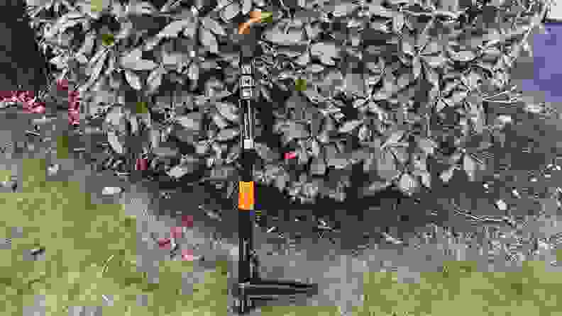 A Fiskars Deluxe Stand-up Weeder leaning against a large garden shrub.