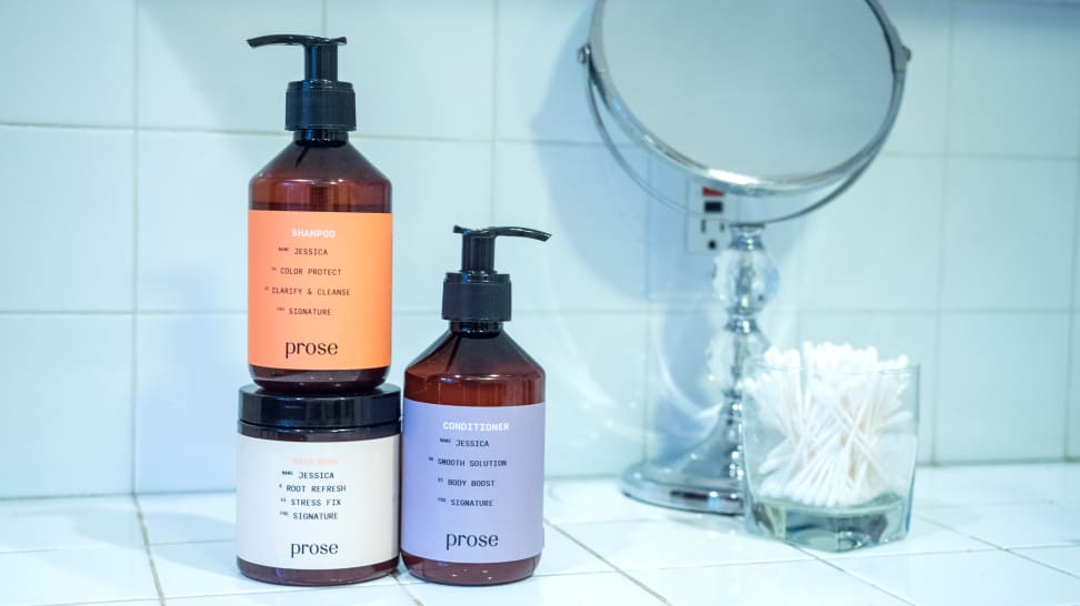 Two pump bottles a jar of hair care sit on a tile vanity next to a container of cotton swaps and a mirror.