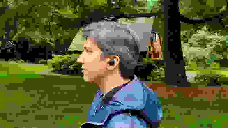 The all-black Sony WF-1000XM4 are shown in a man's ear in front of a park building in a lush green forest.
