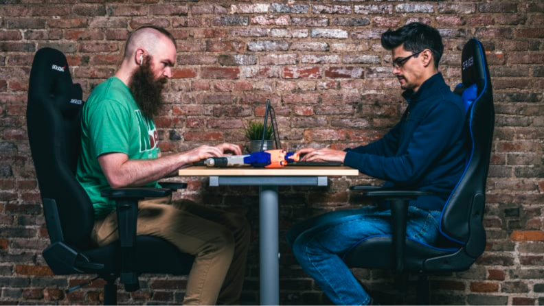 Two people sit at a desk playing video games while sitting in gaming chairs.