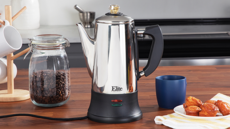 Elite maxi electric coffee percolator on a wooden counter, beside a jar of coffee beans and a plate of desserts