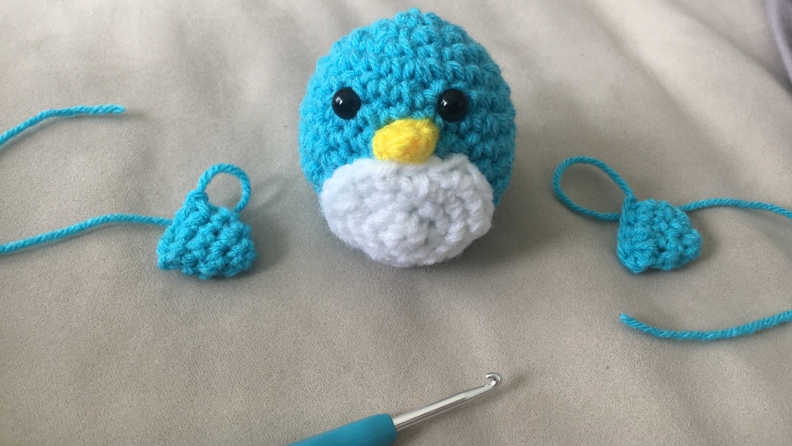 A crocheted penguin sits mid assembly, with his wings off to each side