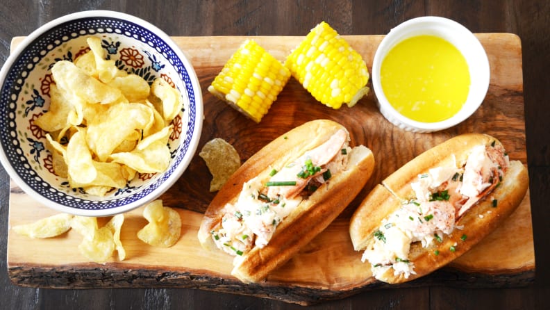 In the center of the image, there's a wooden plate and two lobster rolls on it. Next to the lobster rolls, there's a cup of melted butter, two corns, and a bowl filled with potato chips.