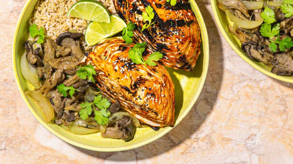 An image of a bowl of rice, mushrooms, and grilled, spicy cabbage on a green plate, garnished with lime wedges.