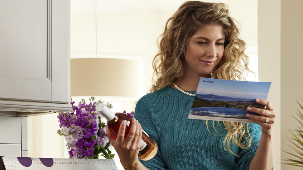A woman in a kitchen holding a bottle of wine and packaging info from a Firstleaf delivery.