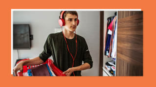 A teenage boy holding a laundry basket inside a bedroom and wearing red corded headphones.