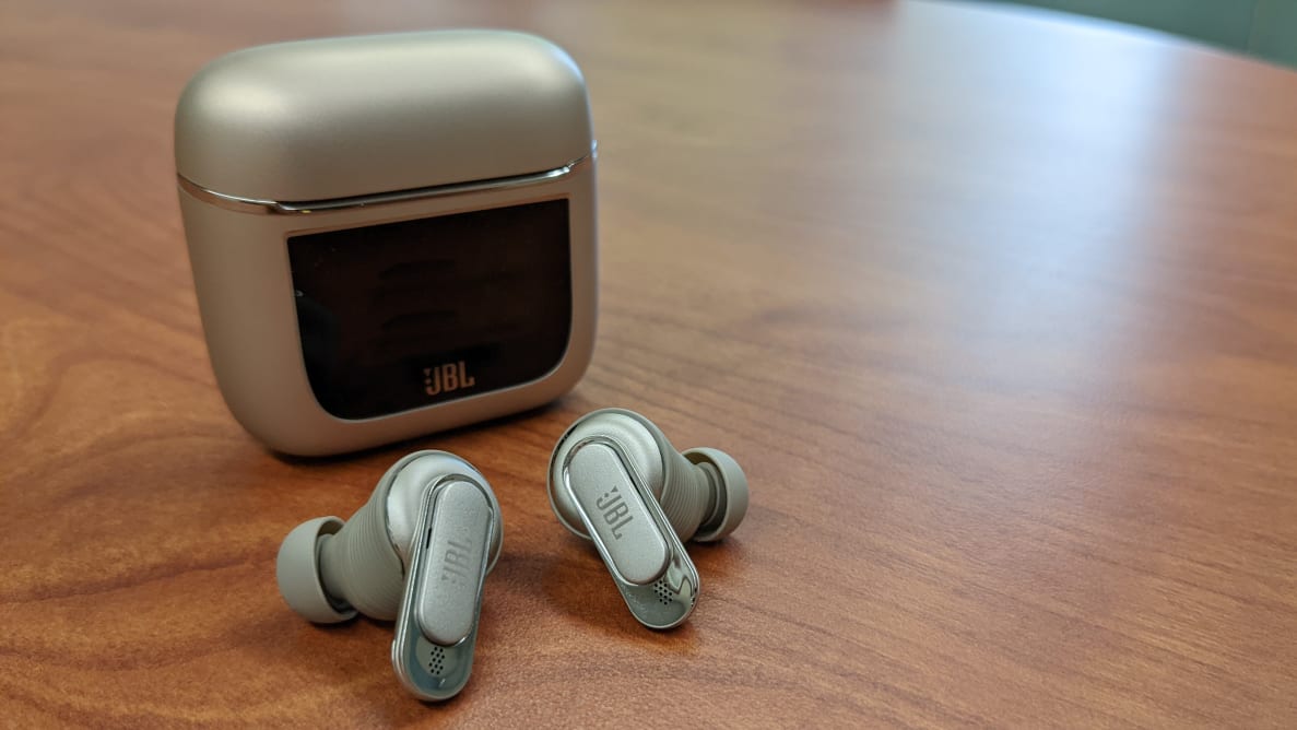 The JBL Tour Pro 2 earbuds sitting in front of their case on a wooden table.