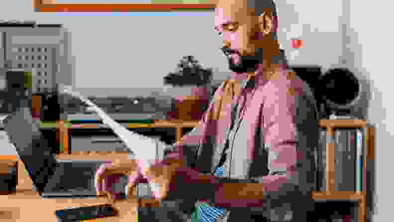 A man sits at a table with a laptop as he holds up financial documents