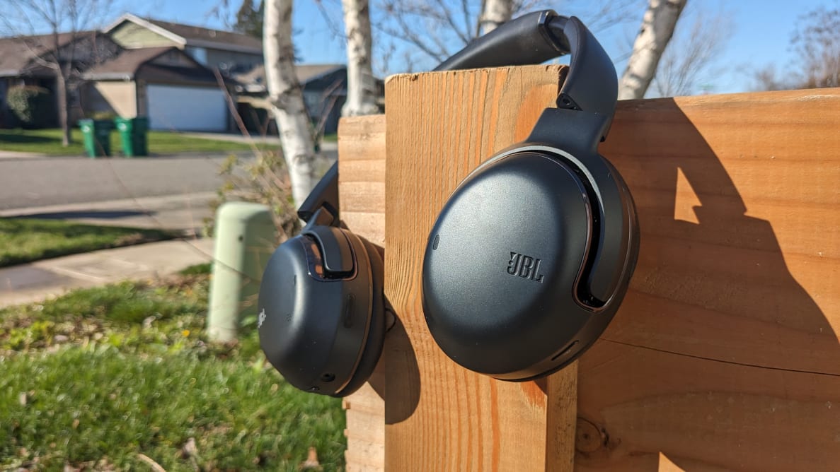 The JBL Tour One M2 headphones hanging on a wooden post on a sunny day.