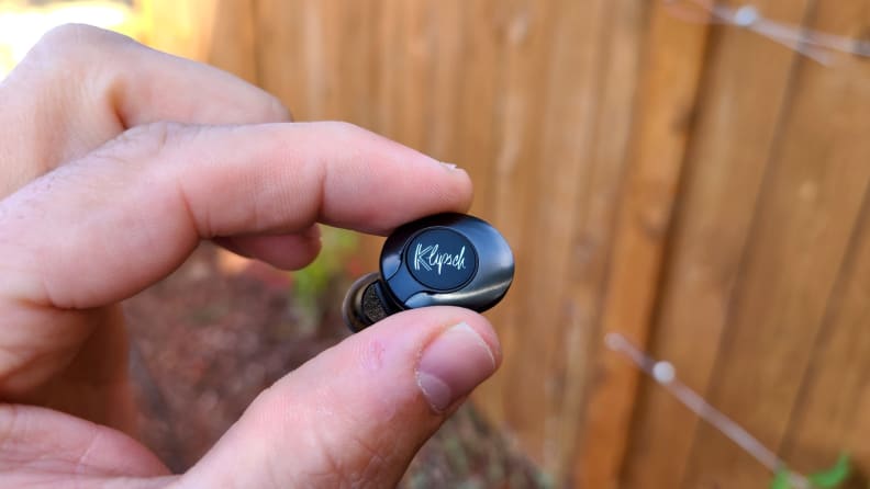 The black enameled Klipsch T5 II True Wireless ANC earbud is held above a lawn and cedar fence, its shiny top glinting in the sun.
