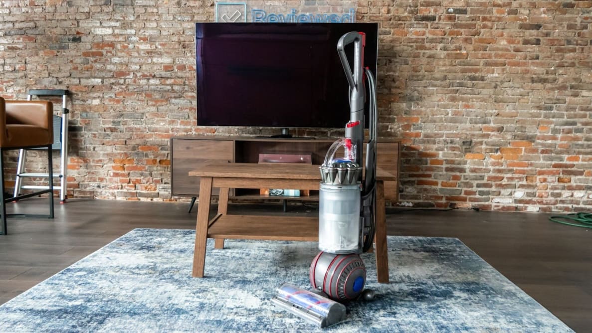 The Dyson Animal 3 in the upright position on blue carpet in front of a television on a wooden stand in front of brick wall.