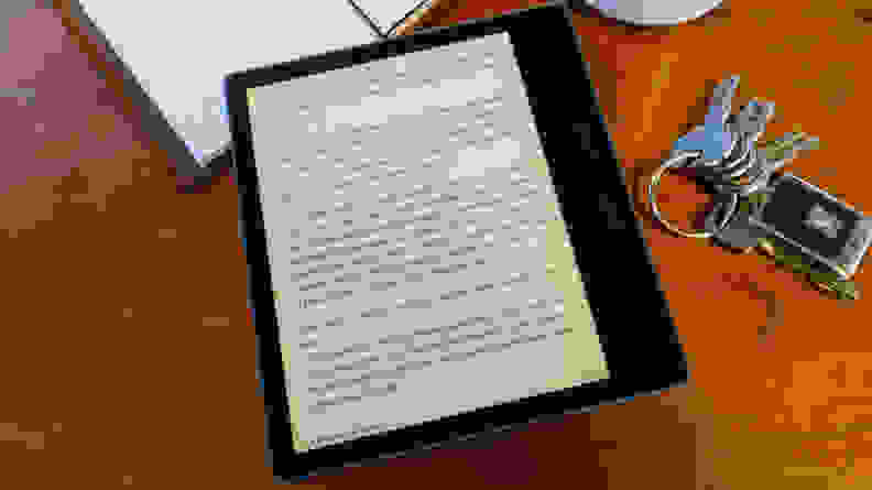A Kindle Oasis (2019) with text from a book on the screen, laid against a wooden table with keys and a notebook in the background.