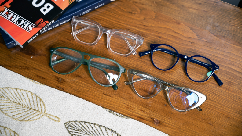 One green, clear, navy and transparent reading glasses laying next to each other on table next to stack of books.