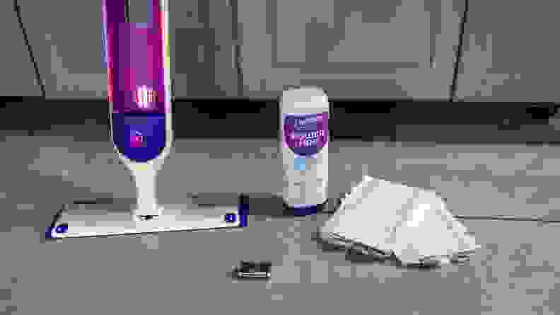 A mop system is positioned next to a bottle of cleaner, mop pads, and parts.