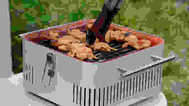 A person is using a pair of tongs to grab a chicken skewer, which is being cooked on an Everdure Cube grill in gray color.