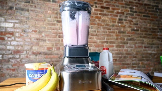 Blender with smoothie next to bananas, yogurt, and other smoothie materials