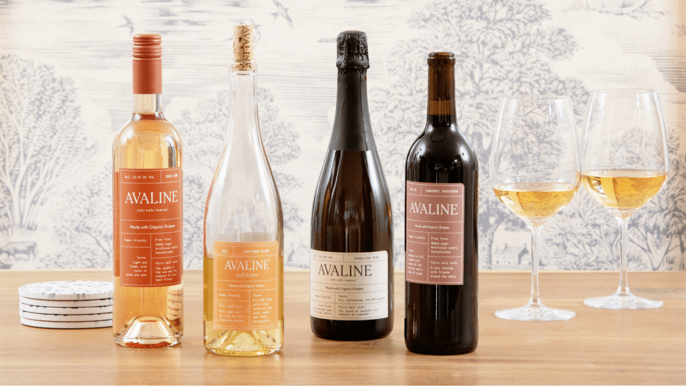 Two white wine and two red wine bottles of Avaline wine and two glasses of white wine on a wood table against a toile background.