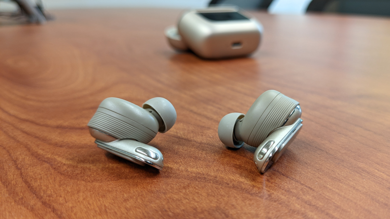 The JBL Tour Pro 2 earbuds sitting on a wooden table with the charging case out of focus in the background.