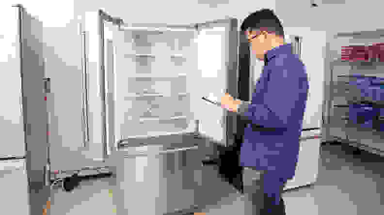 A man stands in front of an open fridge in  with a clipboard.