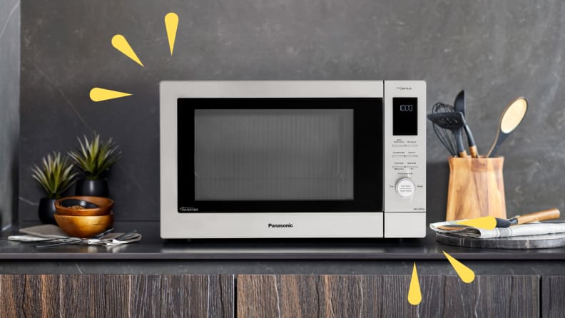 A Panasonic microwave is displayed on a dark kitchen countertop with wooden utensils, small plants, and dishes arranged around it.