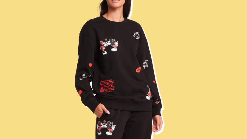 A woman wearing a black crewneck sweatshirt with a picture of Mickey and Minnie Mouse kissing and red and white writing against a yellow background.