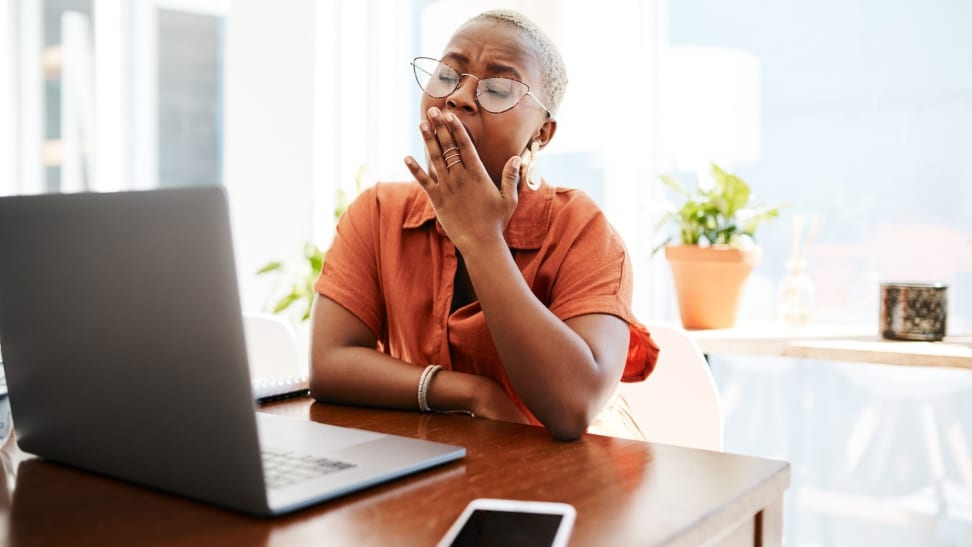 Person yawns with hand over mouth in front of laptop at desk.