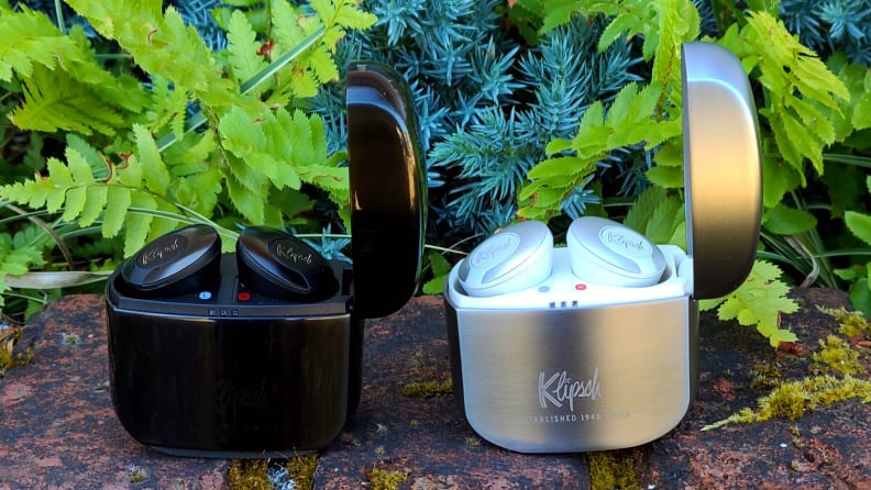 The black enameled Klipsch T5 II True Wireless ANC sit with their metallic case open on a brick ledge next to the silver T5 II earbuds in white, both set in front of greenery.