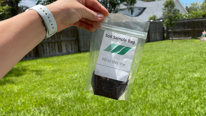A bag of dirt is held up against a backdrop of green grass and a weathered fence.