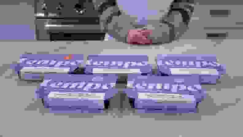 Five pre-packaged Tempo meals inside of individual containers on top of countertop surface in front of person with their hands folded together.