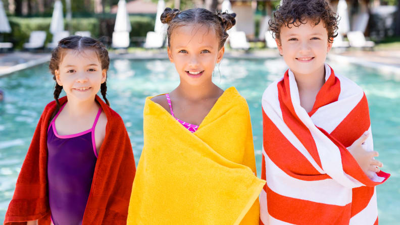 Three young children wrapped in beach towels standing by a pool smiling