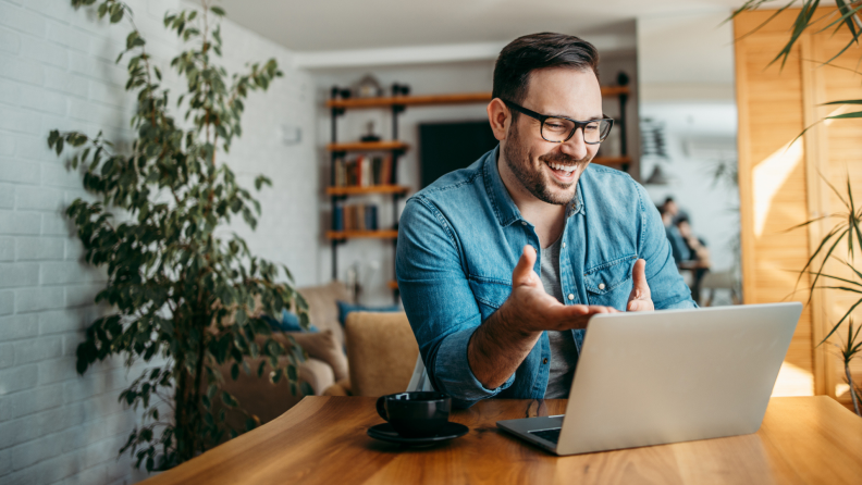 An individual sporting glasses, stubble, and a denim overshirt gestures with their hands, speaking into a laptop. A teacup sits on the table beside the computer; plants and bookshelves are visible in the backdrop.