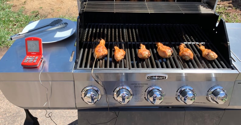 Six chicken drumsticks cooking on a gas grill, one of them hooked up to a probe thermometer