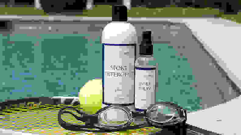 A bottle of The Laundress detergent, a bottle of The Laundress Sport Spray, a tennis ball, and a pair of swimming goggles sitting on a tennis racket in front of a swimming pool.