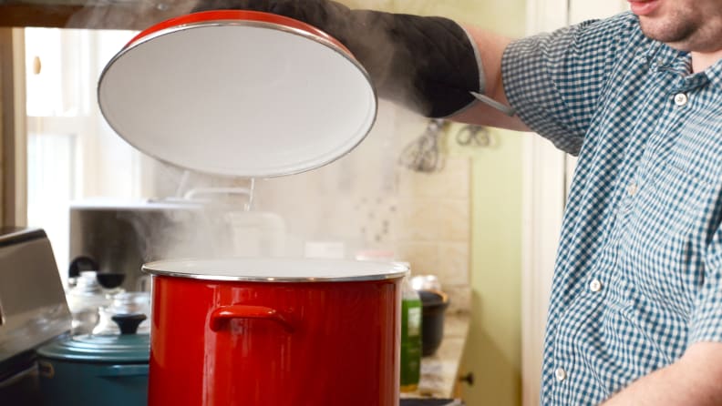 A person is lifting up the lid of a large red stock pot with his mittens on.