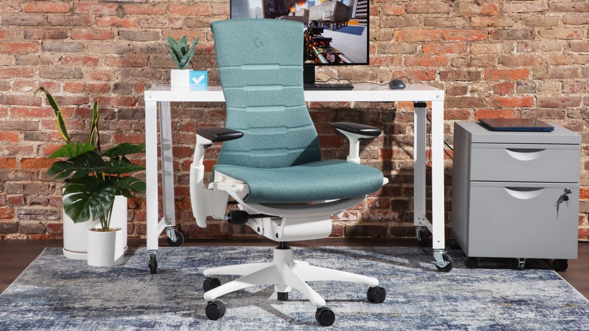The Herman Miller x Logitech G Embody gaming chair, next to a white desk with a monitor and office supplies around it.