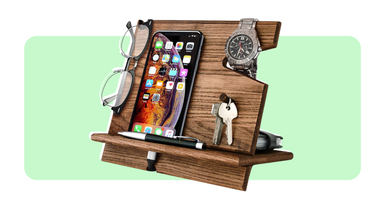 A smartphone, a wristwatch, a set of keys and a pair of glasses sitting on top of wooden dock station in front of light green background.