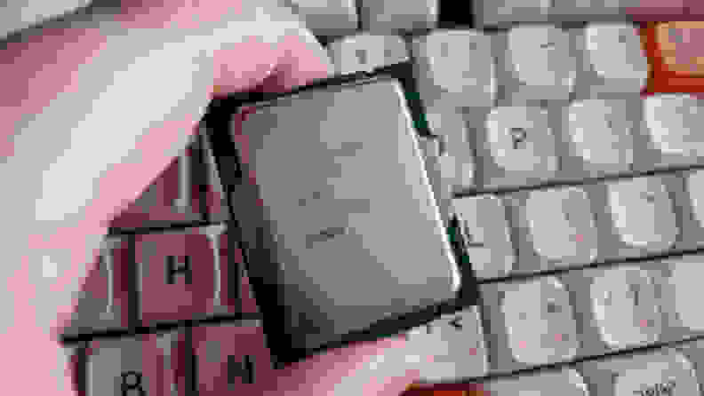 A desktop computer processor grapsed between two fingers with a white keyboard in the background.