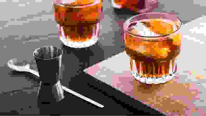 A pair of amber-colored cocktails with orange wedges sit on a table beside a cocktail jigger.
