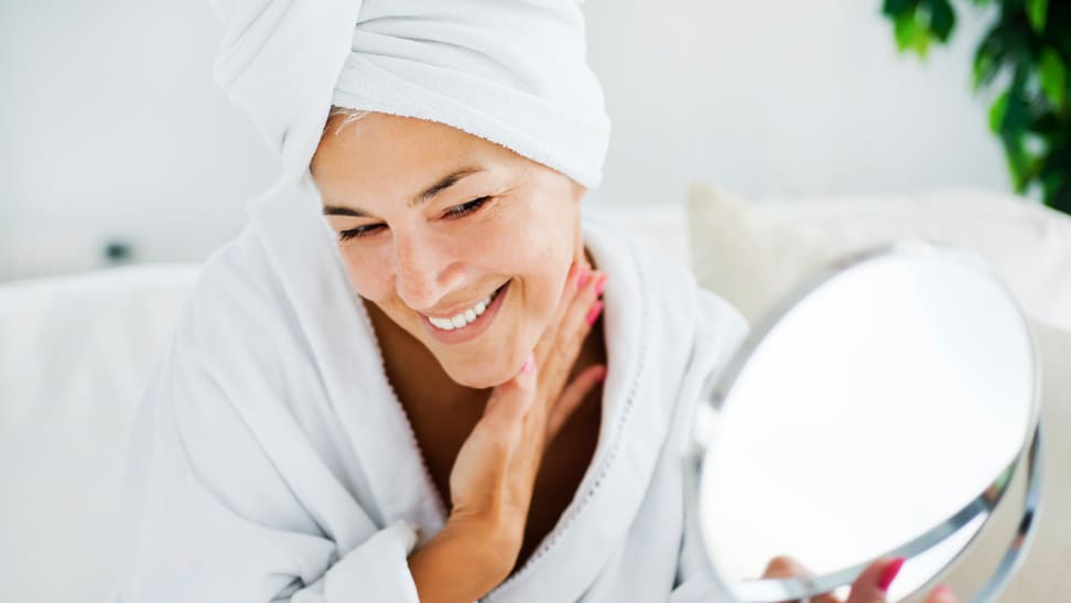 A person wearing a robe and a towel on their head examining their face in a mirror.