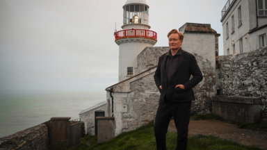 An image of Conan O'Brien standing on a shoreline alongside a lighthouse.