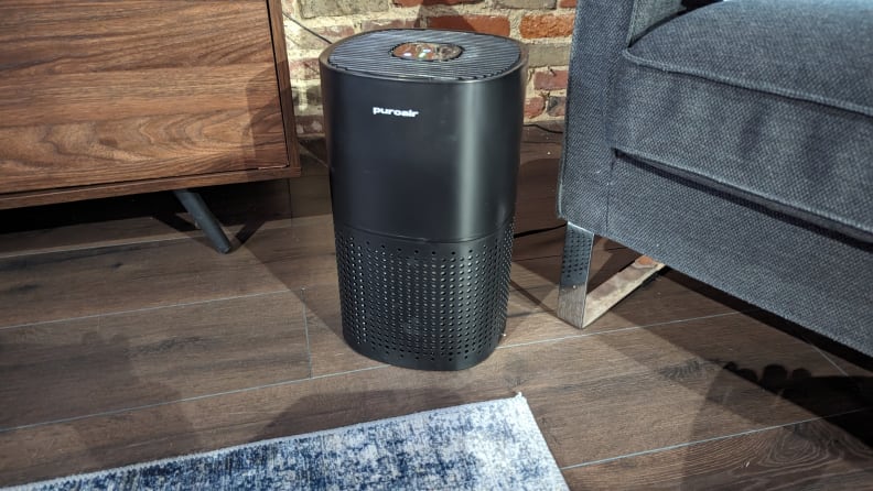 A black  Puro Air 240 air purifier stands aside a gray couch and on a brown wood floor.