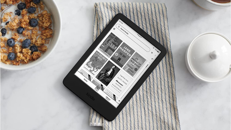 An Amazon Kindle resting on a blanket on a bed next to a bowl of cereal and a bowl of sugar.