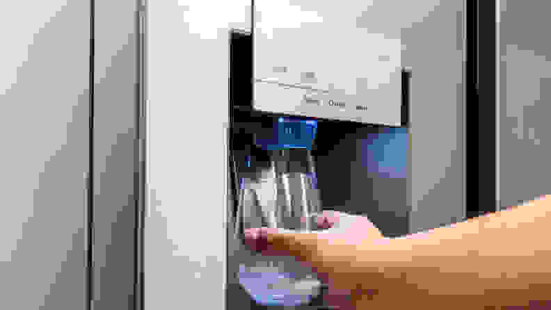 A hand reaches into frame holding a glass against the through-the-door dispenser's paddle. The fridge is dispensing water into the glass.