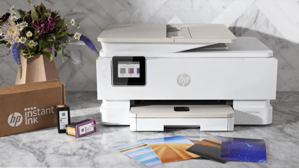 An image of a white HP Envy printer next to a spread of photos on a table. An HP Instant Ink delivery box sits off to the left of the printer.