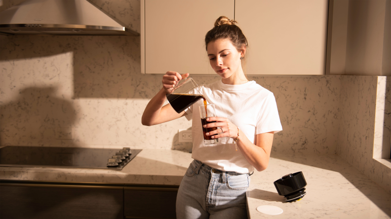 A person wearing a white shirt is pouring coffee from a glass carafe into a cup in the kitchen.