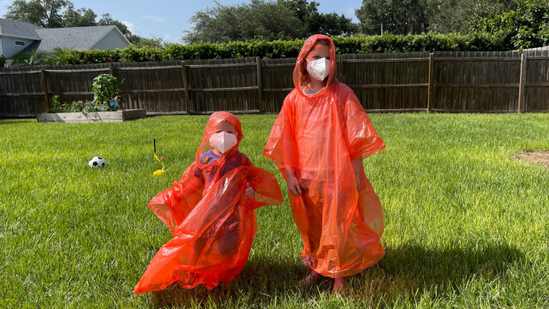 Two kids standing outside in the grass wearing orange ponchos and white face masks.