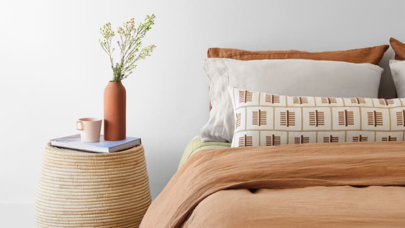 Bed with tan comforter and patterned pillows next to woven side table with a plant sitting on top